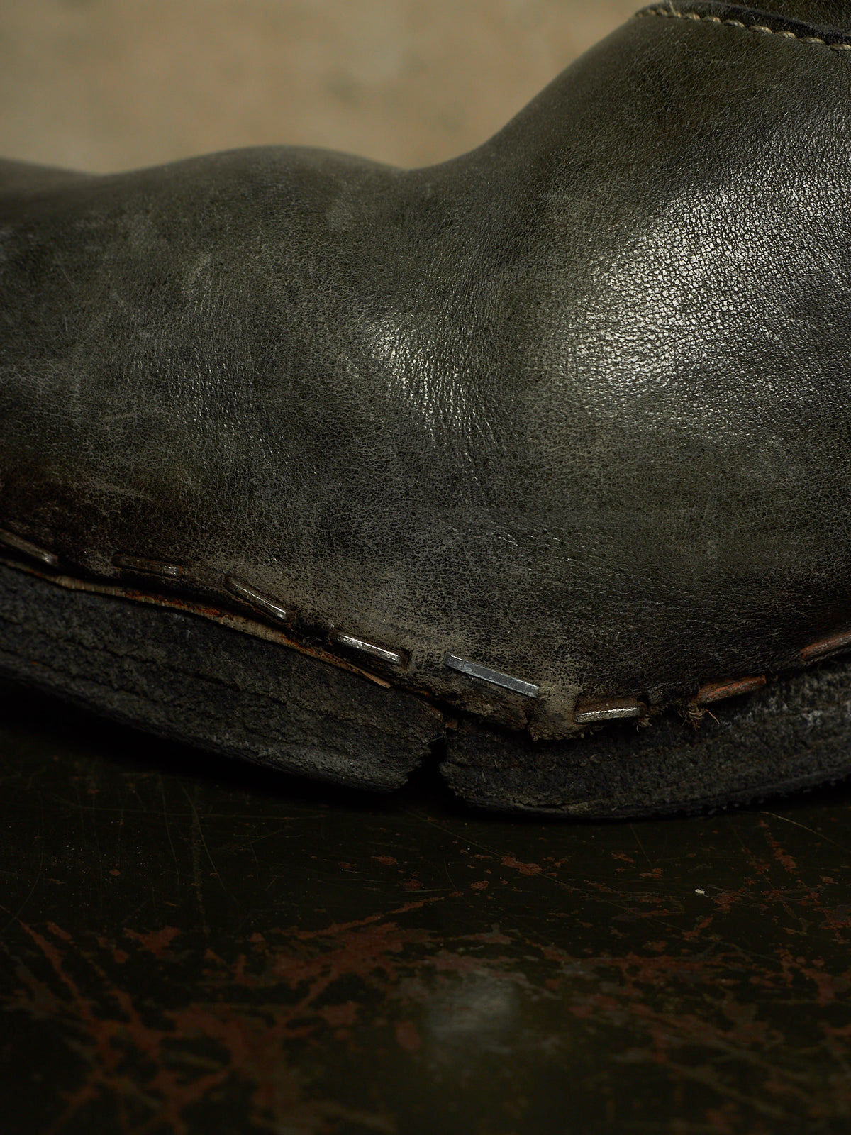 Close-up of a dark distressed leather boot showing the side profile, raw stitching, and sole edge.