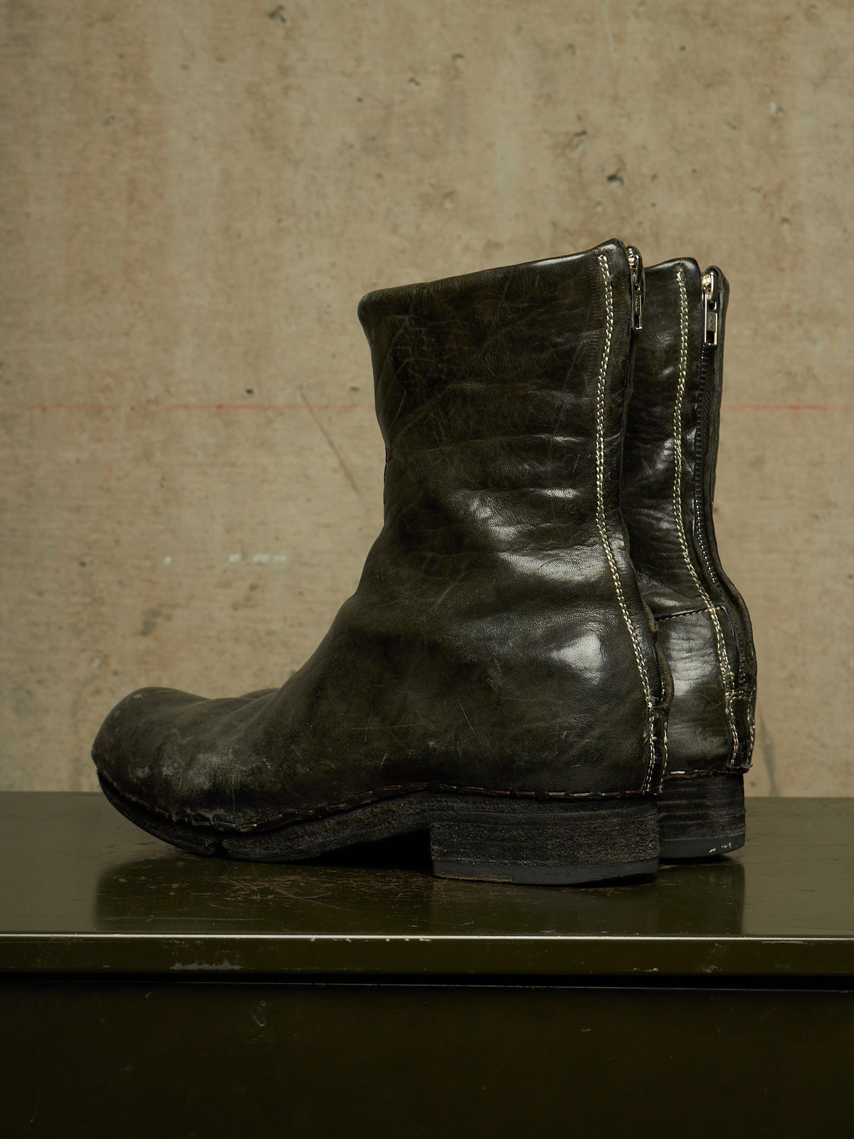A single dark olive green leather ankle boot with side zippers, shown in profile against a textured beige concrete wall.