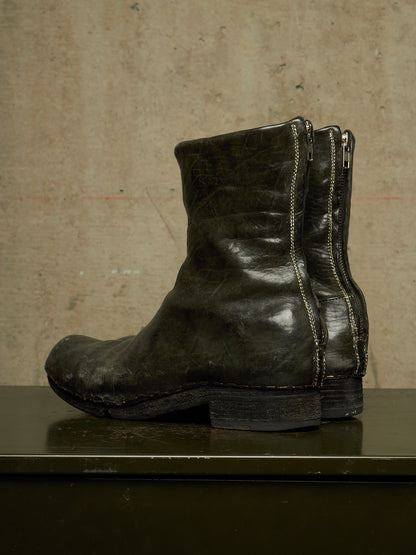 A single dark olive green leather ankle boot with side zippers, shown in profile against a textured beige concrete wall.
