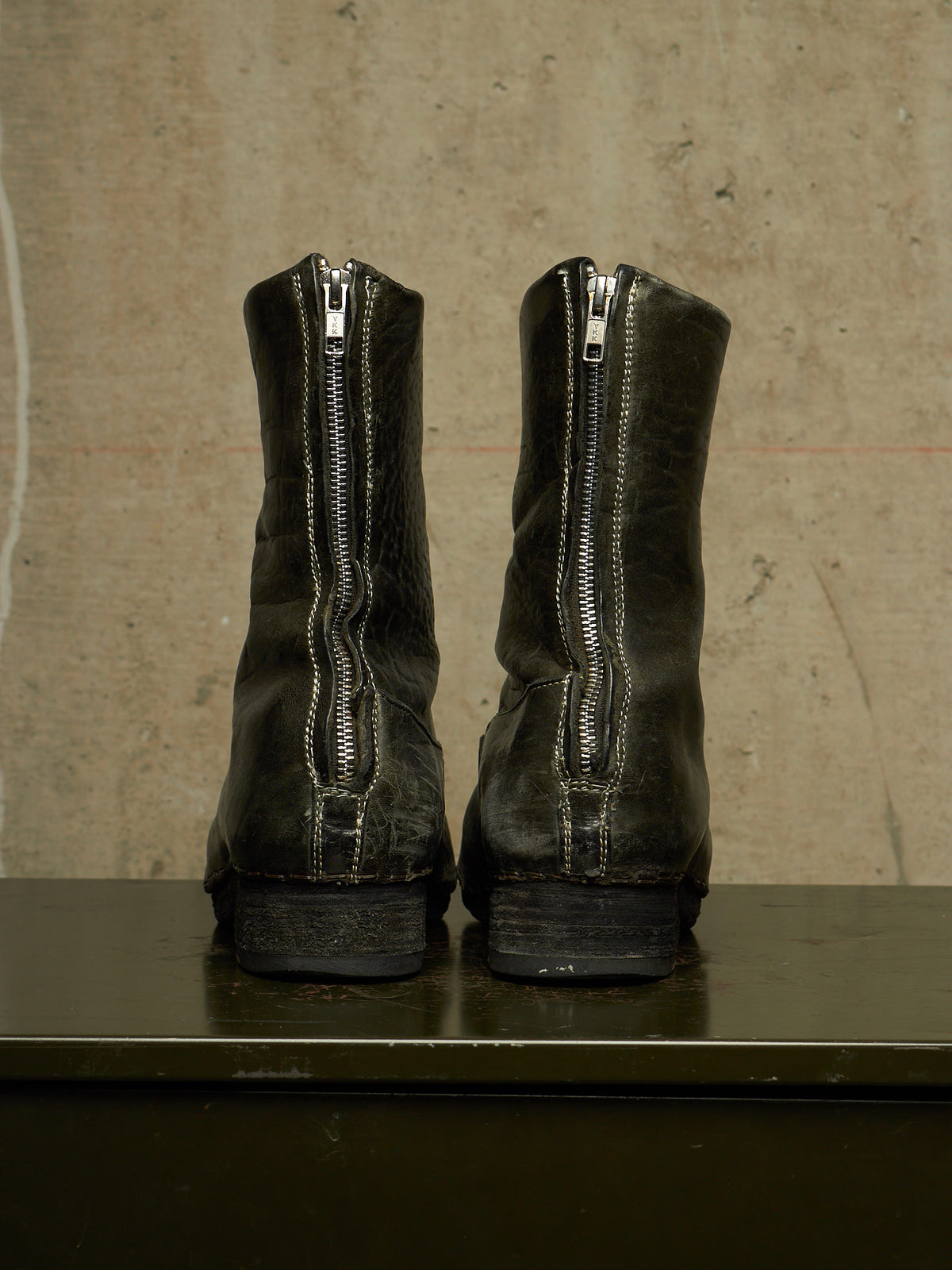 A pair of black leather ankle boots with silver zippers on the back, shown from the rear on a dark surface against a textured concrete wall.