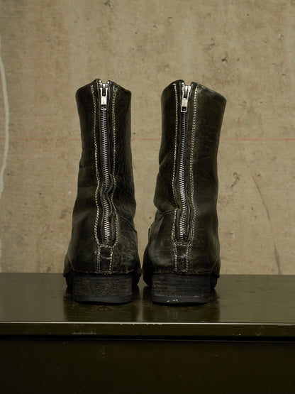 A pair of black leather ankle boots with silver zippers on the back, shown from the rear on a dark surface against a textured concrete wall.