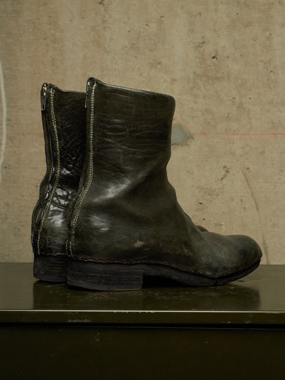 A pair of dark olive green leather ankle boots with side zippers, displayed on a dark green surface against a textured beige wall.