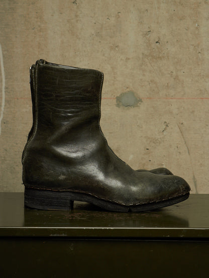 A single dark olive-green leather ankle boot with a low block heel and side zipper, resting on a dark surface against a textured beige wall.