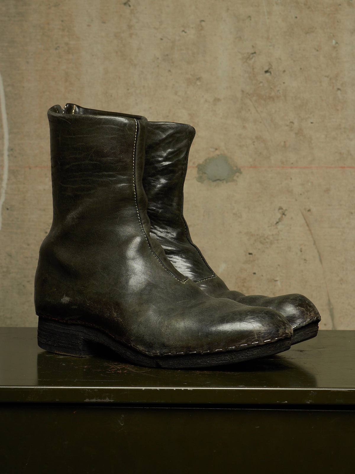 A single dark olive green leather ankle boot with a distressed finish, resting on a dark green surface against a textured beige wall.