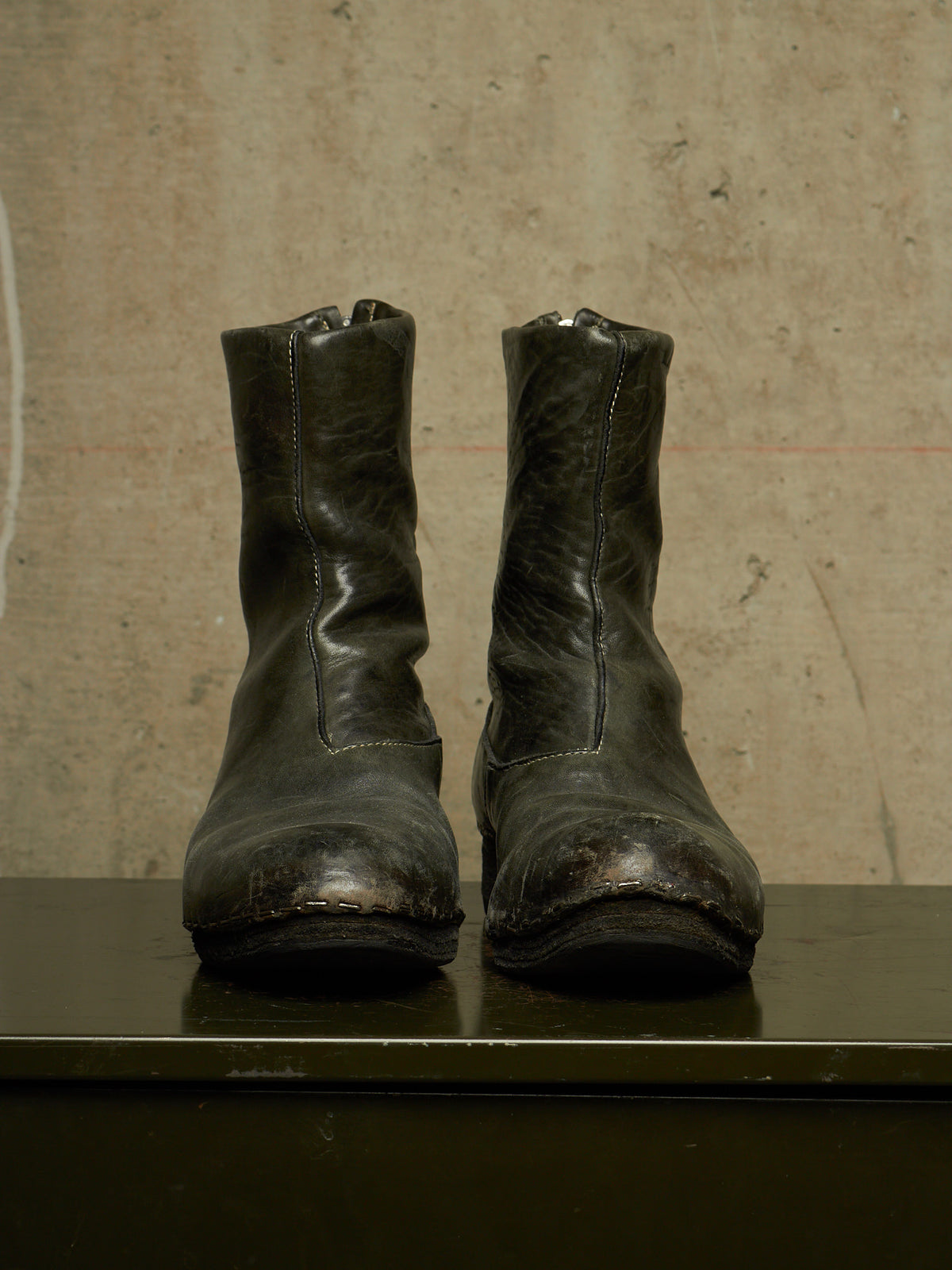 A pair of worn black leather ankle boots with distressed detailing, displayed side-by-side on a dark surface against a textured concrete wall.