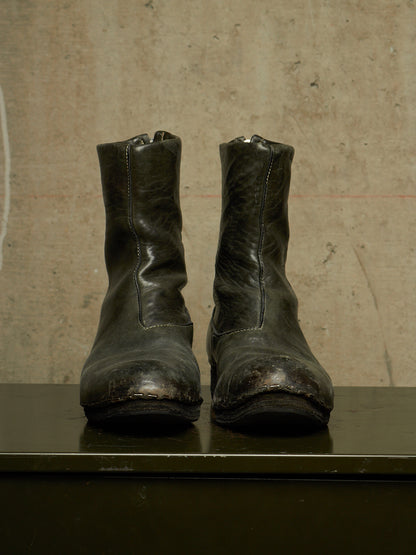 A pair of worn black leather ankle boots with distressed detailing, displayed side-by-side on a dark surface against a textured concrete wall.