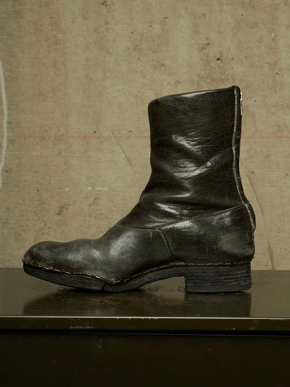 A single dark olive green leather ankle boot with a low stacked heel, shown in profile against a textured beige concrete wall.