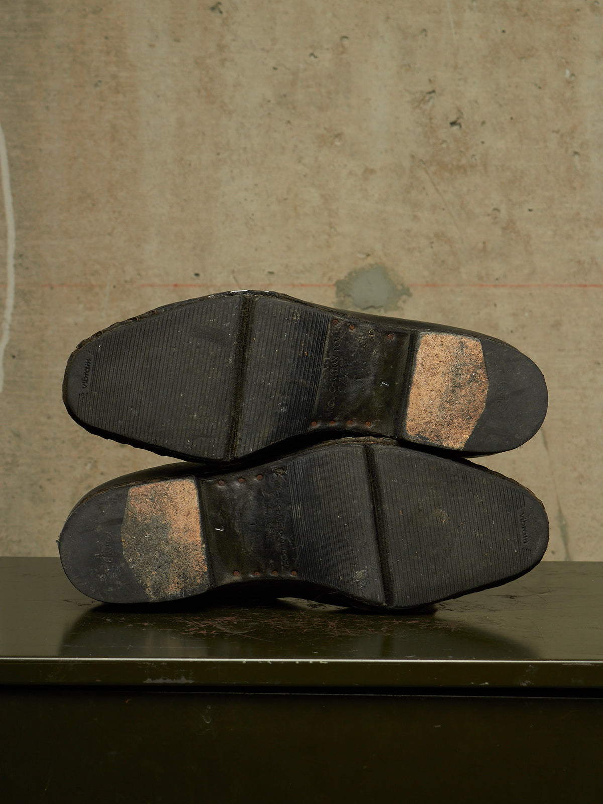 A pair of black rubber shoe soles viewed from underneath, resting on a dark surface against a textured beige wall.