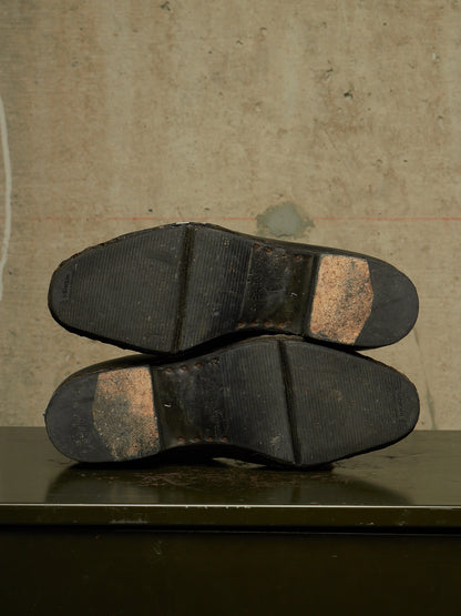A pair of black rubber shoe soles viewed from underneath, resting on a dark surface against a textured beige wall.