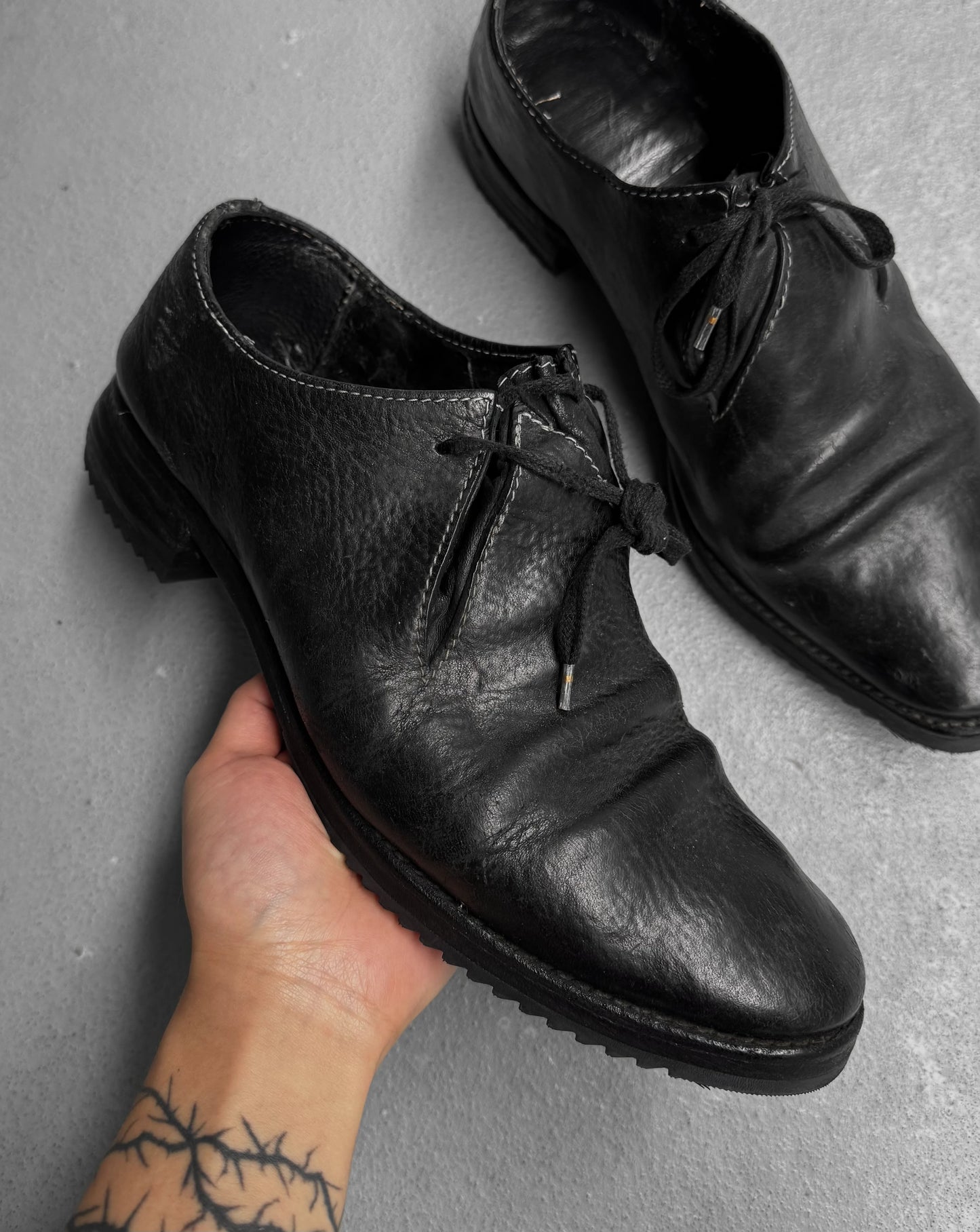 Black leather shoes on a gray surface with a hand holding one shoe.