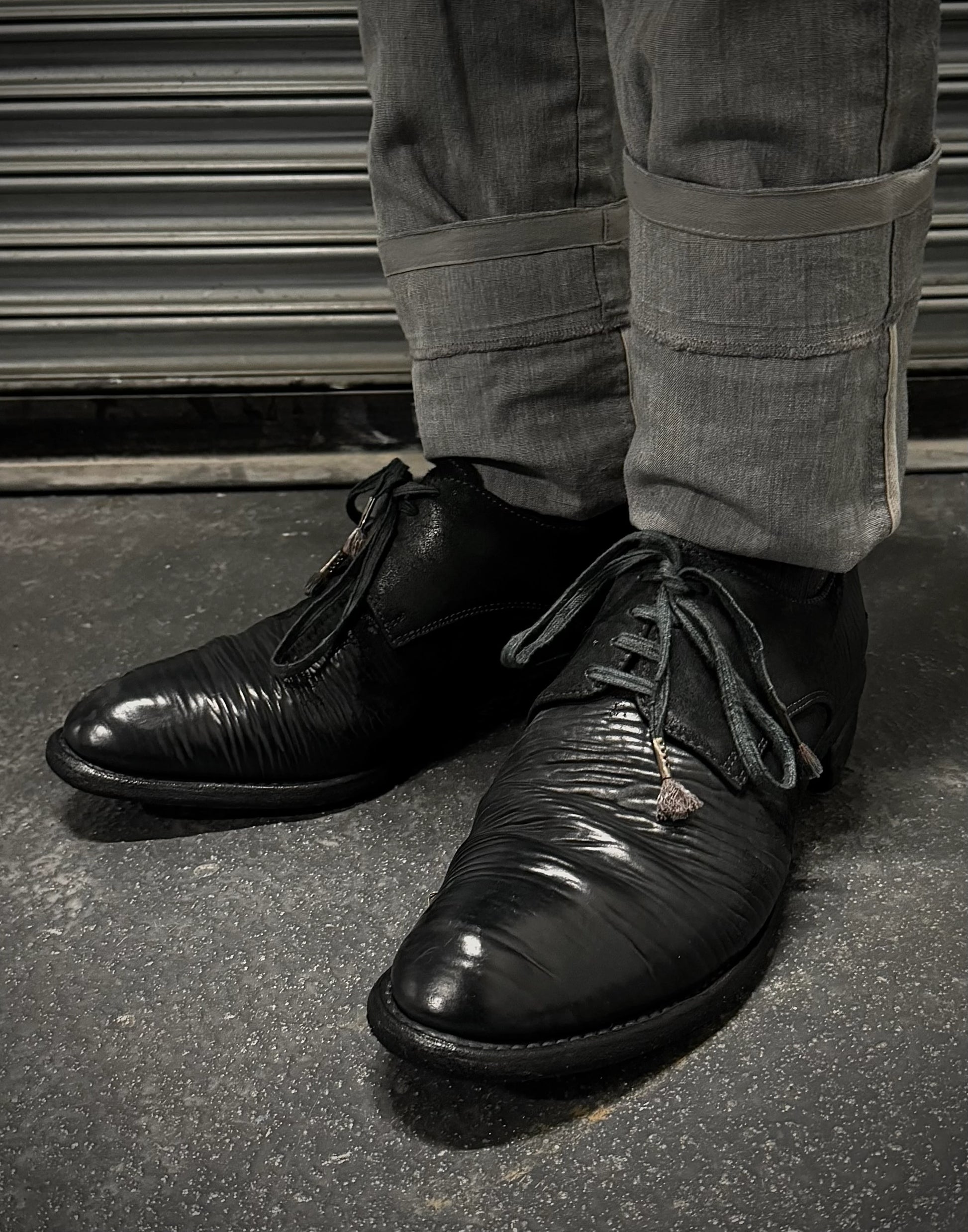 Black leather shoes worn with gray pants on a dark surface.