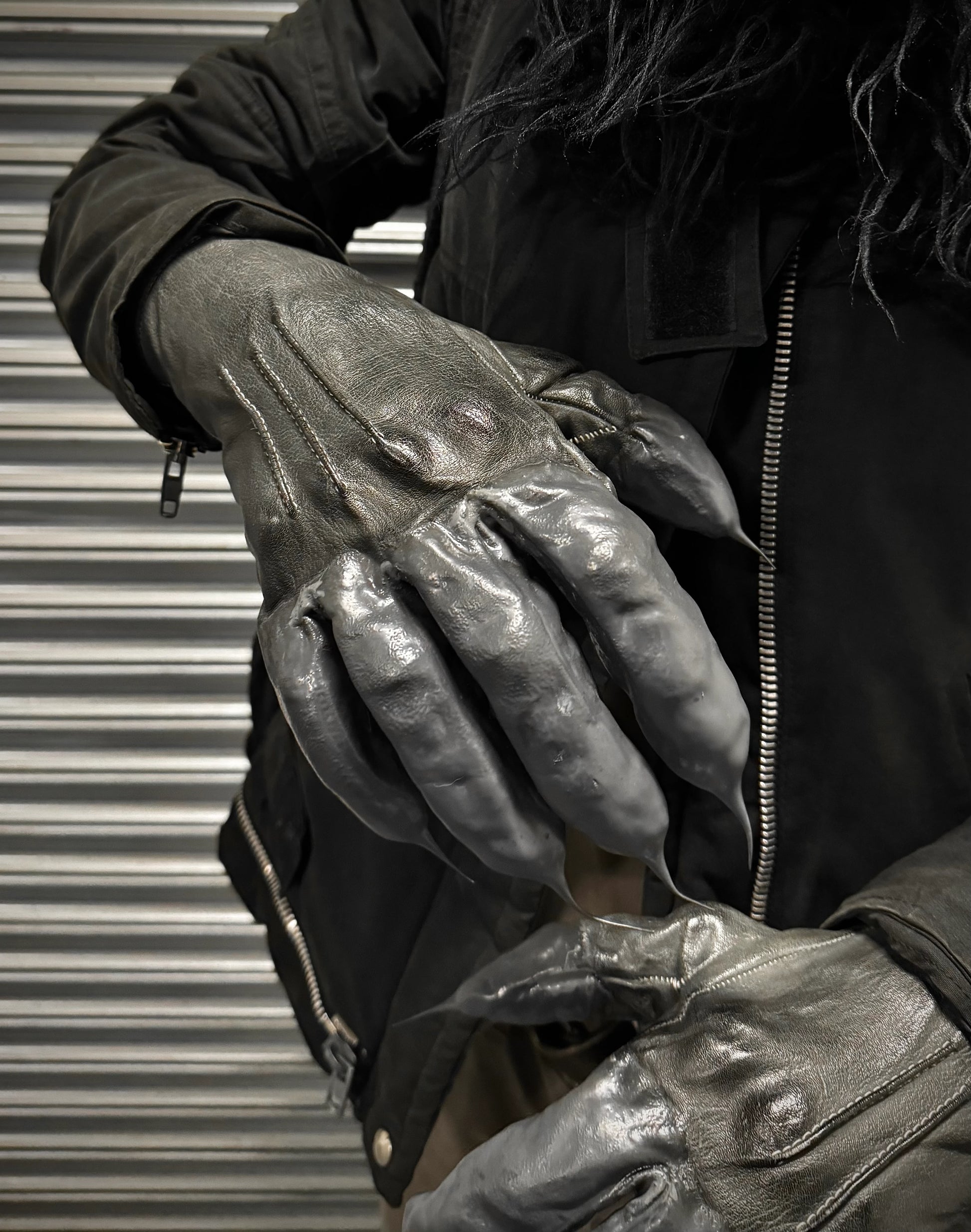 Person wearing shiny leather gloves with a metallic sheen against a corrugated metal background.