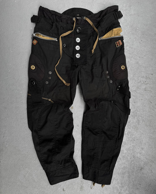 The L.G.B Early 00s Pilot Cargo Capris by L.G.B are black capris with multiple zippers, buttons, and pockets, accented with brown and tan fabrics and inspired by Japanese fashion label aesthetics, shown laid flat on a light gray surface.