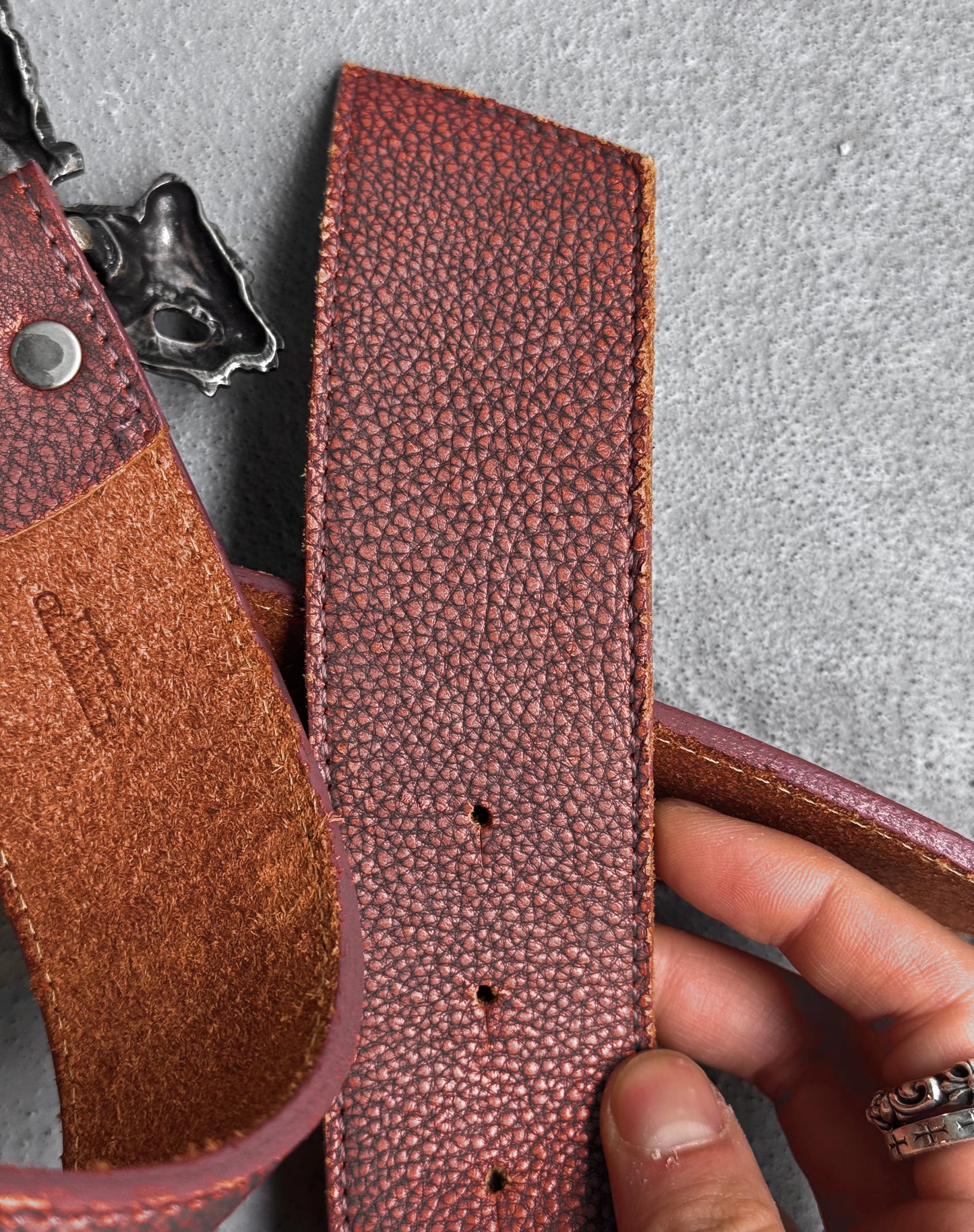 A person holds a Tornado Mart Early 00s Pirate Silver Buckle Leather Belt, size M (US 32-36 Waist), in flawless textured brown leather with silver buckle and rivets, displayed on a gray surface.