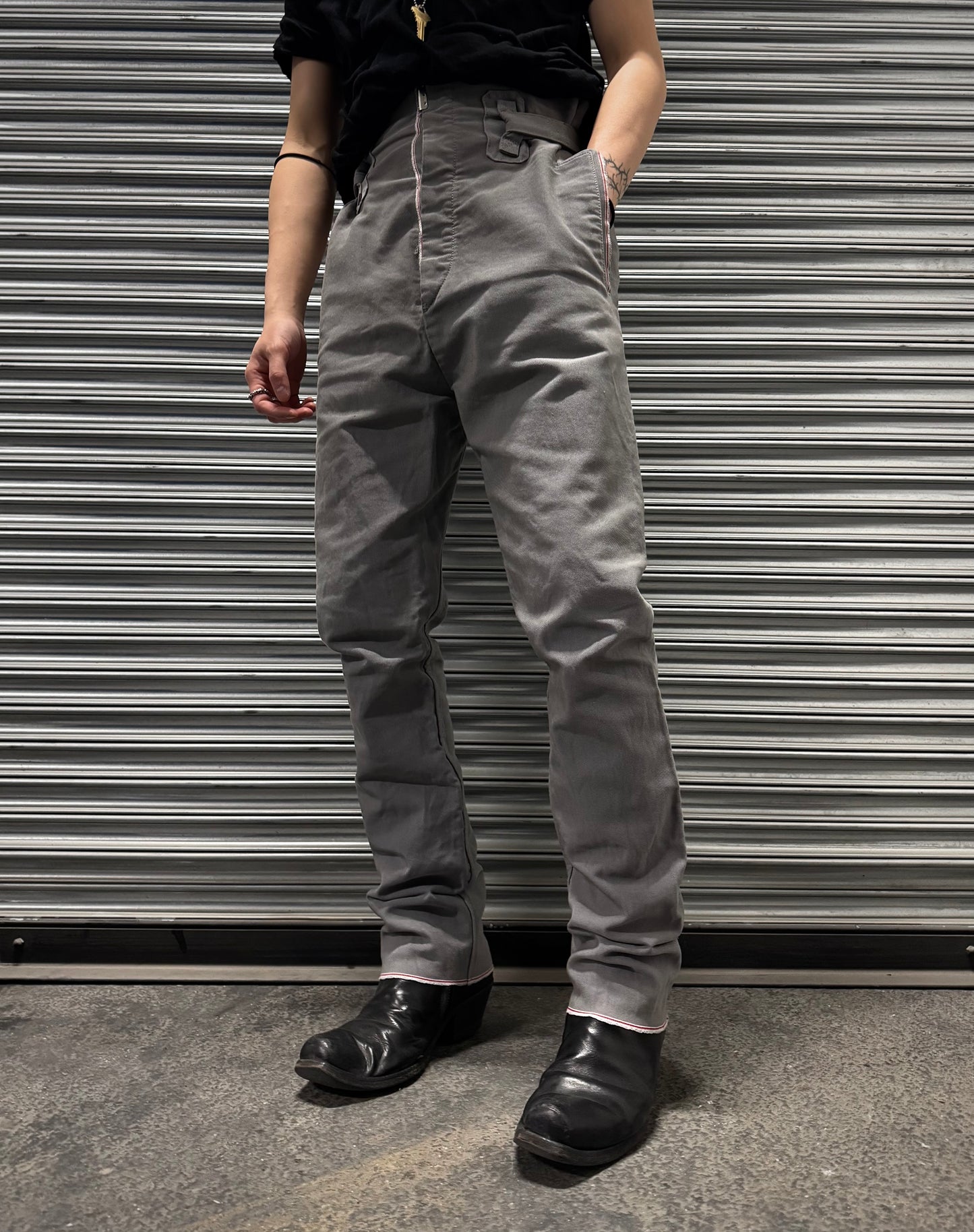 A person stands before a metal shutter, sporting avant-garde style in a black shirt, black boots, and Carol Christian Poell 2010 “IN-BETWEEN” PM/2578 PEDGE self-edge high-waisted metre pants. Their head is out of frame and arms rest naturally at their sides.