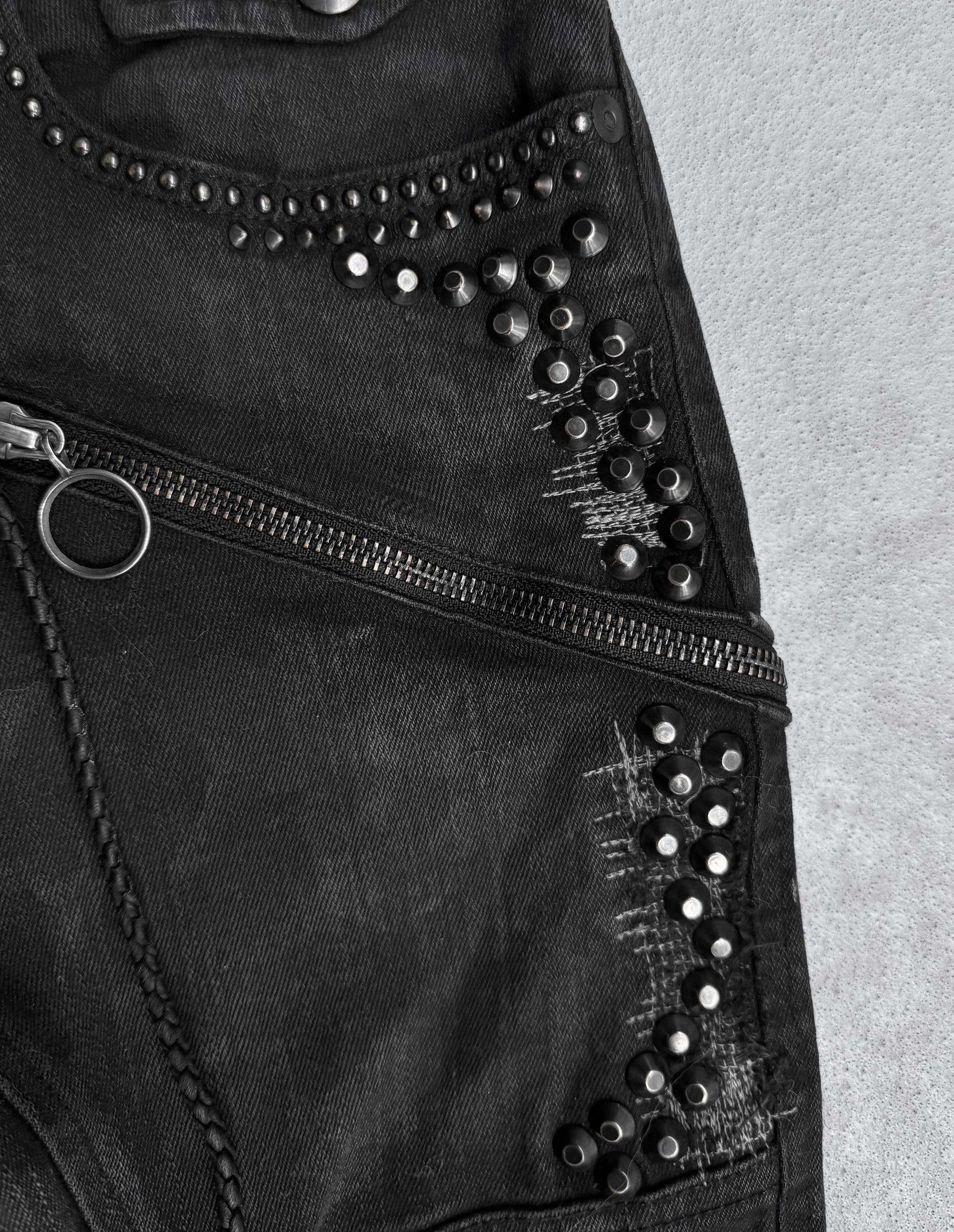 Close-up of KMRii Early 00s Goatskin Patches Studded Crush Flare-zip Denim: black denim with fading, diagonal zipper, decorative studs, distressed details. Textured surface in black and white. Fits US 29-30 waist or size 2.