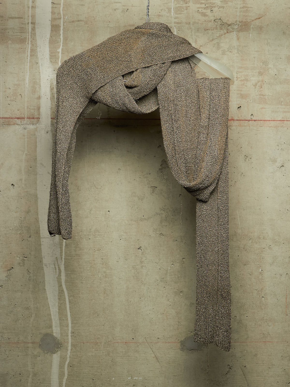 A textured, chunky knit scarf in a heathered taupe-brown color, draped and wrapped around a white hanger against a concrete wall.