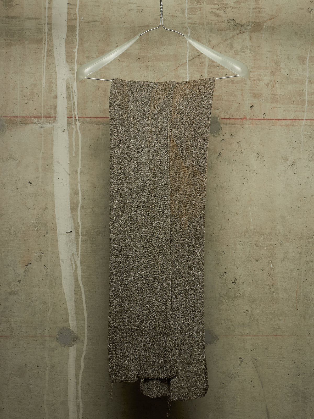 A textured, heathered taupe-brown knit garment or fabric panel hanging from a clear plastic hanger against a concrete wall.