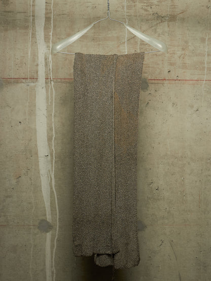 A textured, heathered taupe-brown knit garment or fabric panel hanging from a clear plastic hanger against a concrete wall.