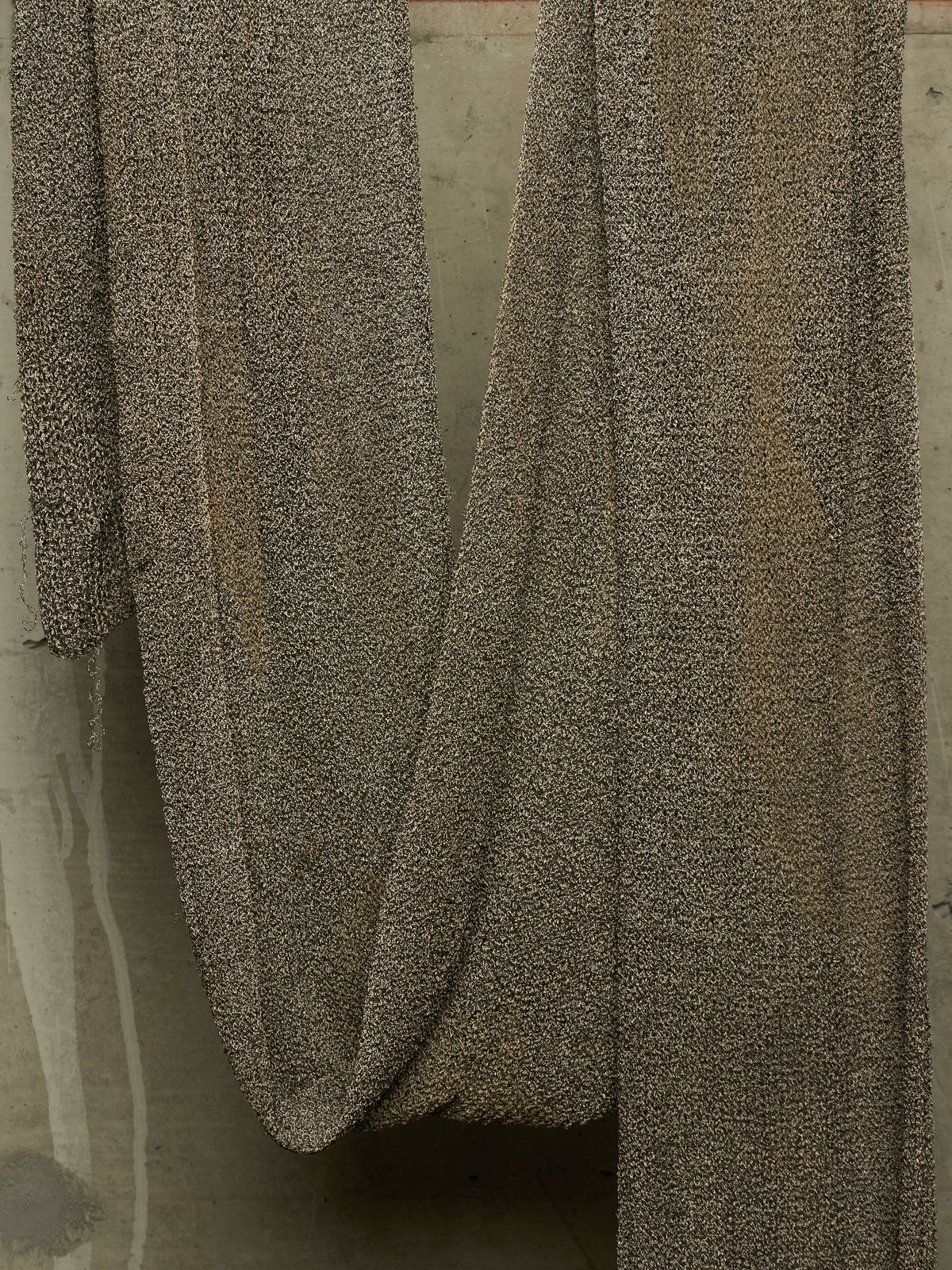 A close-up of draped textured fabric in a speckled taupe and brown pattern, hanging against a neutral concrete-like background.