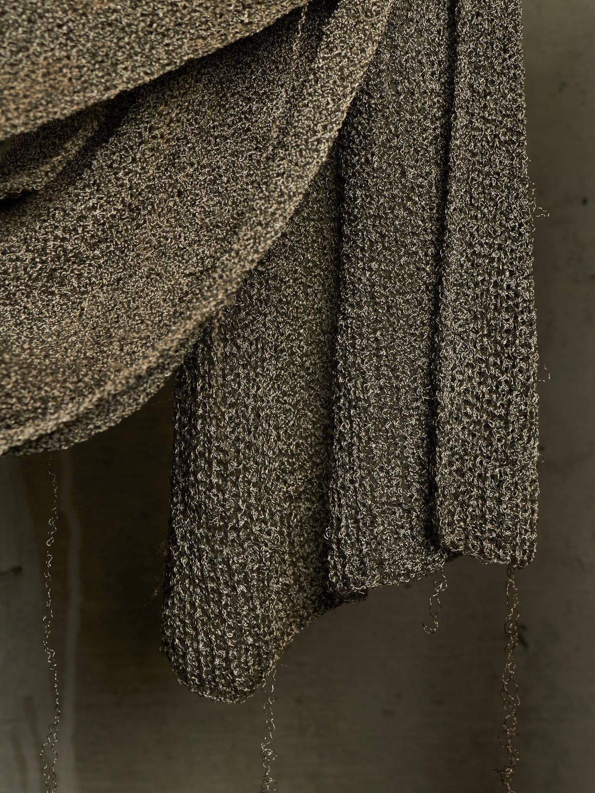 A close-up of a draped, chunky knit textile in a dark taupe-brown color with subtle metallic flecks, hanging against a neutral gray background.