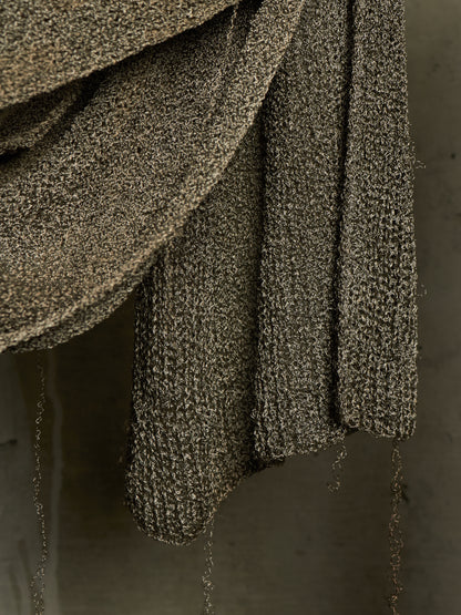 A close-up of a draped, chunky knit textile in a dark taupe-brown color with subtle metallic flecks, hanging against a neutral gray background.