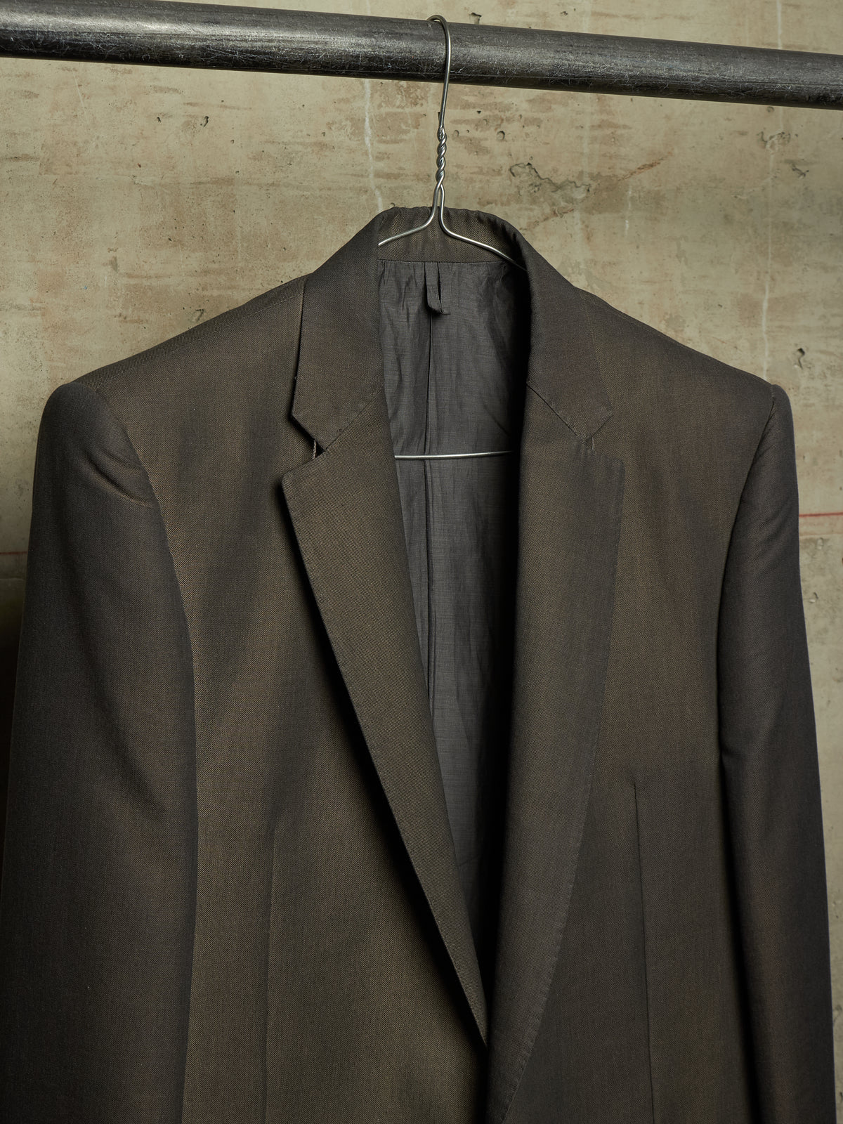 A dark brown tailored suit jacket hanging on a metal hanger against a textured concrete wall.