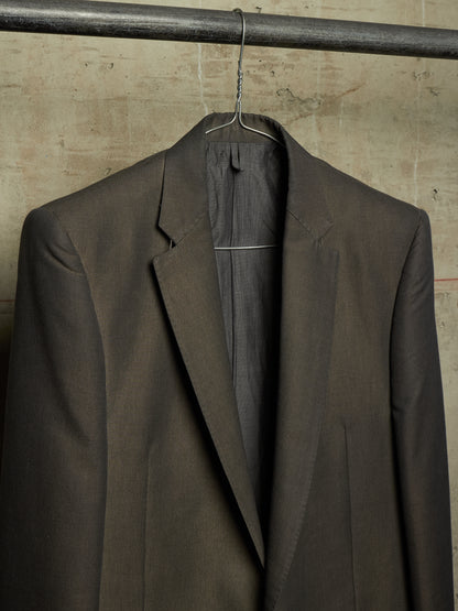A dark brown tailored suit jacket hanging on a metal hanger against a textured concrete wall.