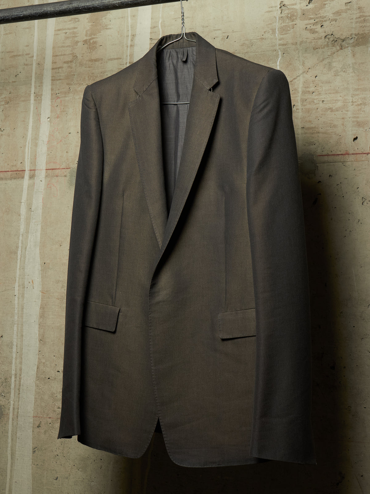 A dark brown tailored suit jacket hanging on a hanger against a textured concrete wall.