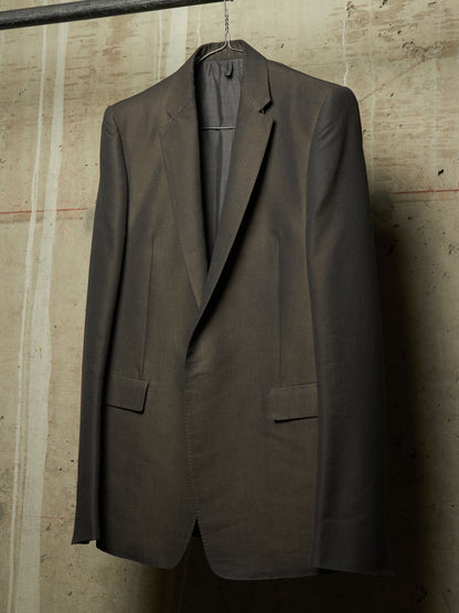 A dark brown tailored suit jacket hanging on a hanger against a textured concrete wall.