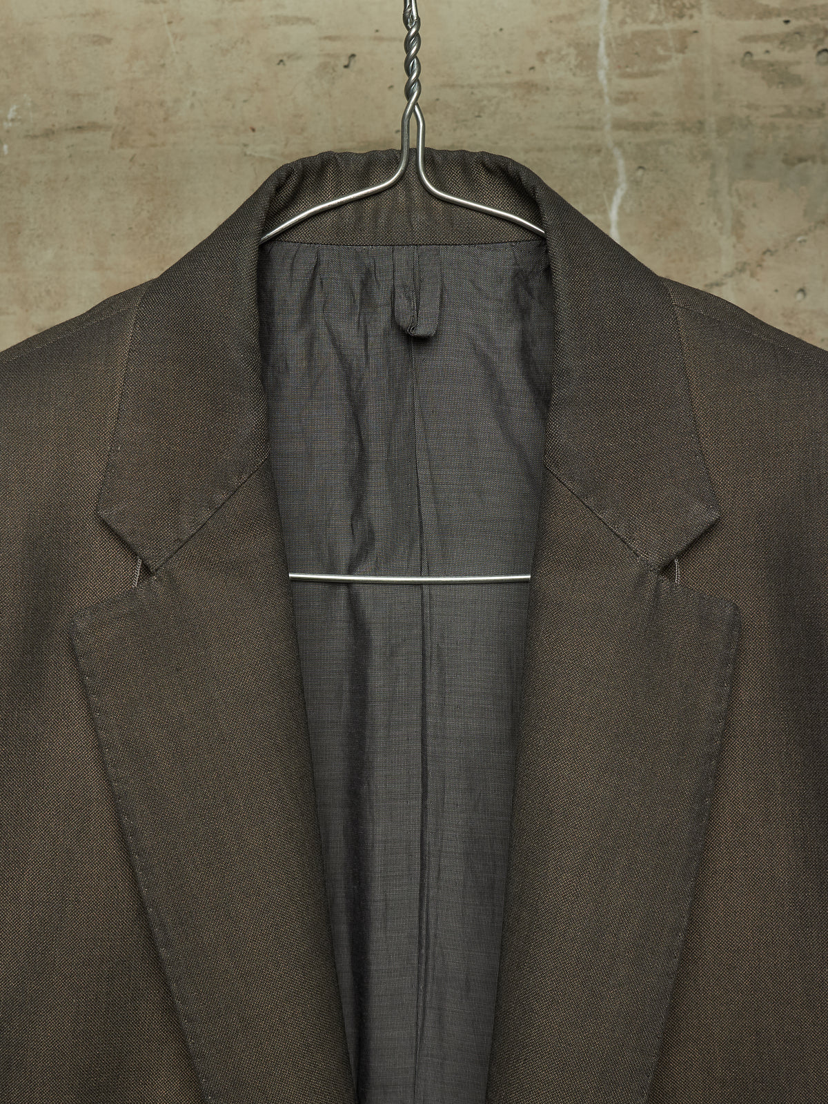 A close-up of a dark brown textured blazer hanging on a metal hanger against a textured beige wall.
