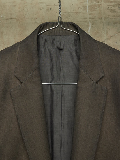 A close-up of a dark brown textured blazer hanging on a metal hanger against a textured beige wall.