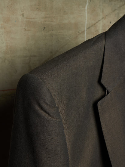 Close-up of a dark brown textured wool suit jacket, showing the shoulder, collar, and lapel against a textured concrete wall background.