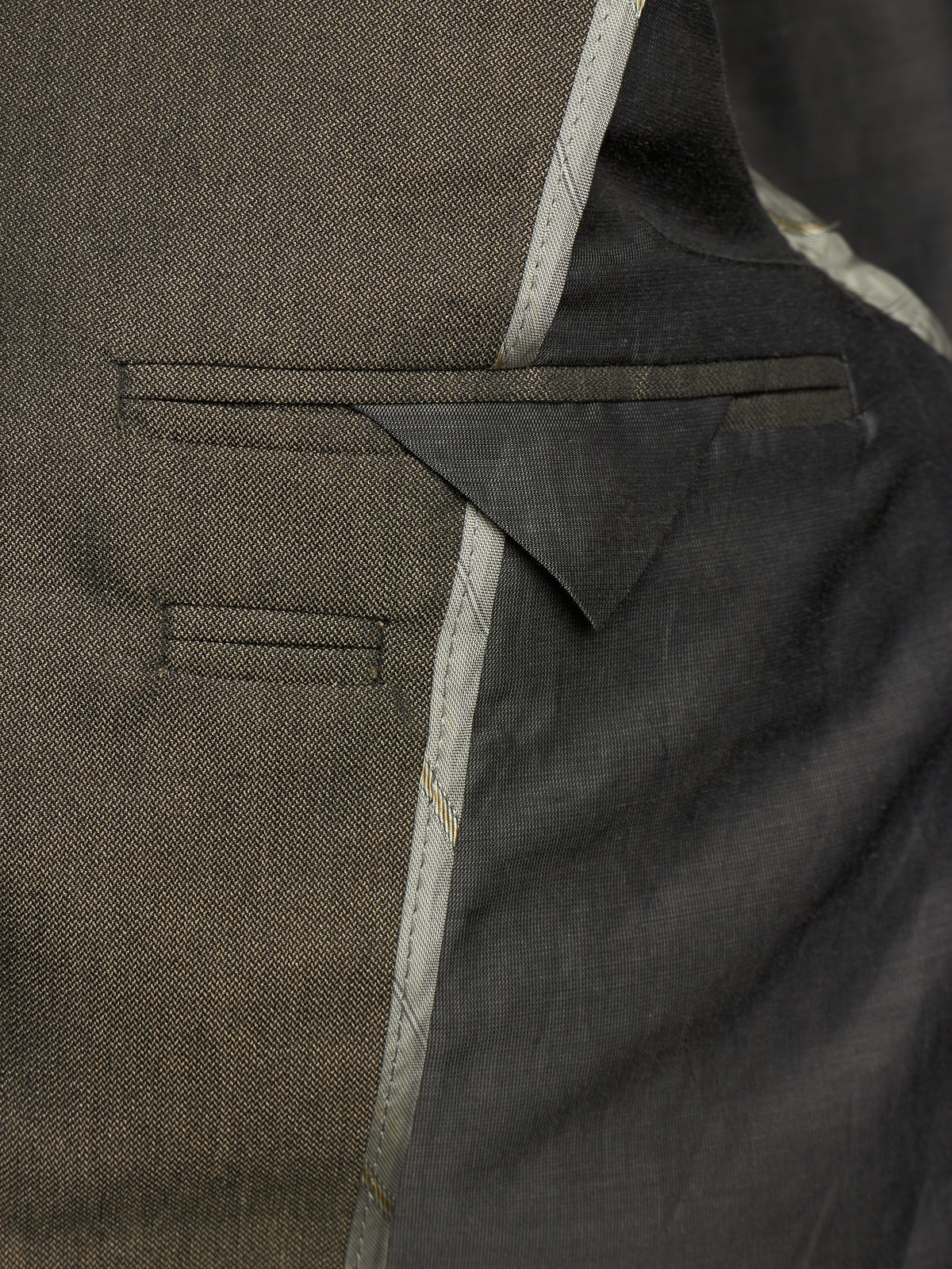 Close-up detail of a tailored jacket or blazer showing a welt pocket, fabric texture, and contrast stitching in dark olive and charcoal tones.
