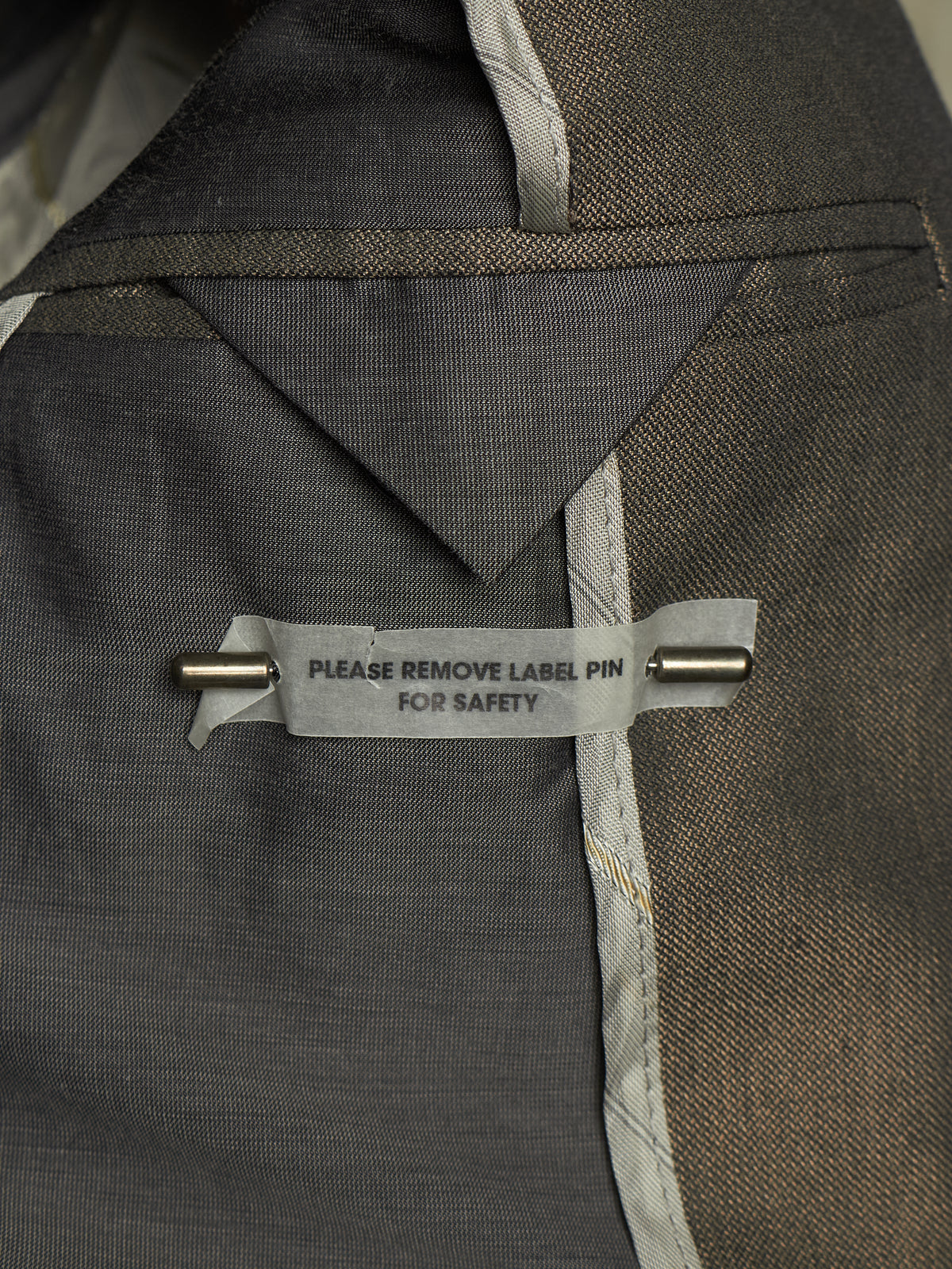 Close-up of a dark gray textured fabric garment with a gray safety label pinned with a metal pin. The label reads 'PLEASE REMOVE LABEL PIN FOR SAFETY'.