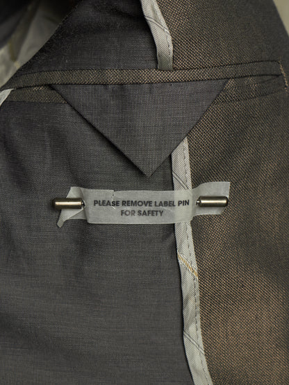 Close-up of a dark gray textured fabric garment with a gray safety label pinned with a metal pin. The label reads 'PLEASE REMOVE LABEL PIN FOR SAFETY'.