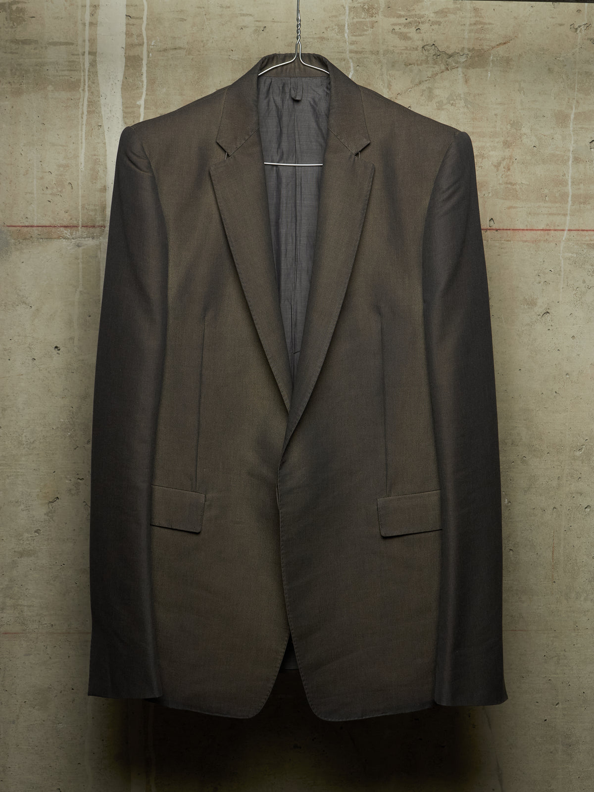 A dark brown tailored suit jacket hanging on a wire hanger against a textured concrete wall.