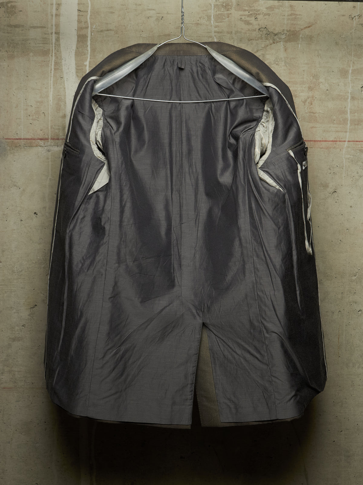 A dark gray waterproof raincoat hanging on a wire hanger against a textured concrete wall. The coat is shown from the back, featuring a high collar, side pockets, and a glossy finish.