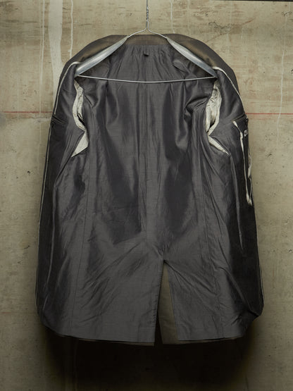 A dark gray waterproof raincoat hanging on a wire hanger against a textured concrete wall. The coat is shown from the back, featuring a high collar, side pockets, and a glossy finish.