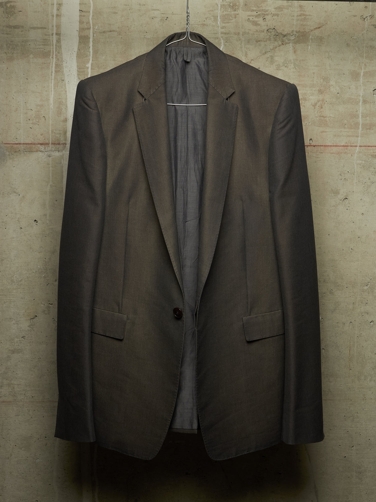 A dark brown tailored blazer jacket hanging on a wire hanger against a textured concrete wall.