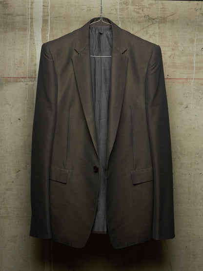 A dark brown tailored blazer jacket hanging on a wire hanger against a textured concrete wall.