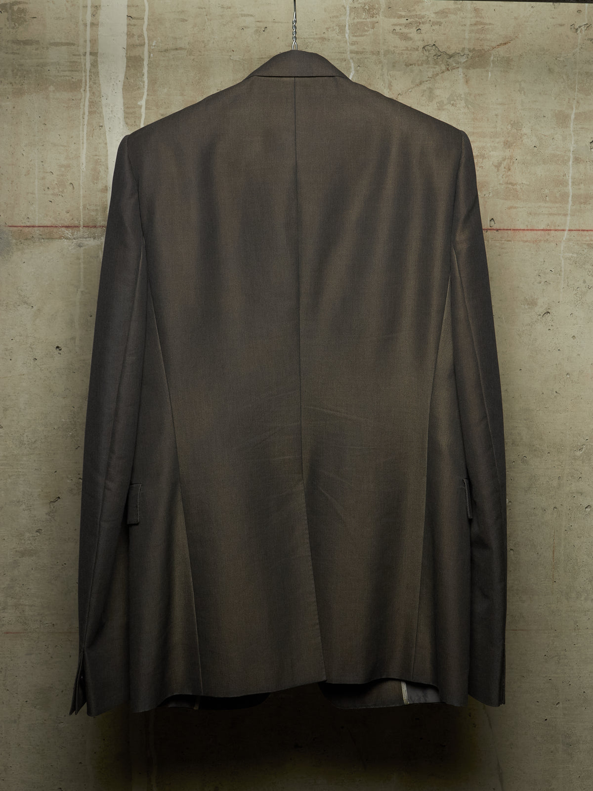 A dark brown tailored suit jacket hanging on a hanger against a textured concrete wall, showing the back view.