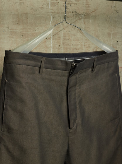 A pair of dark olive gray trousers hanging on a clear plastic hanger against a textured concrete wall.