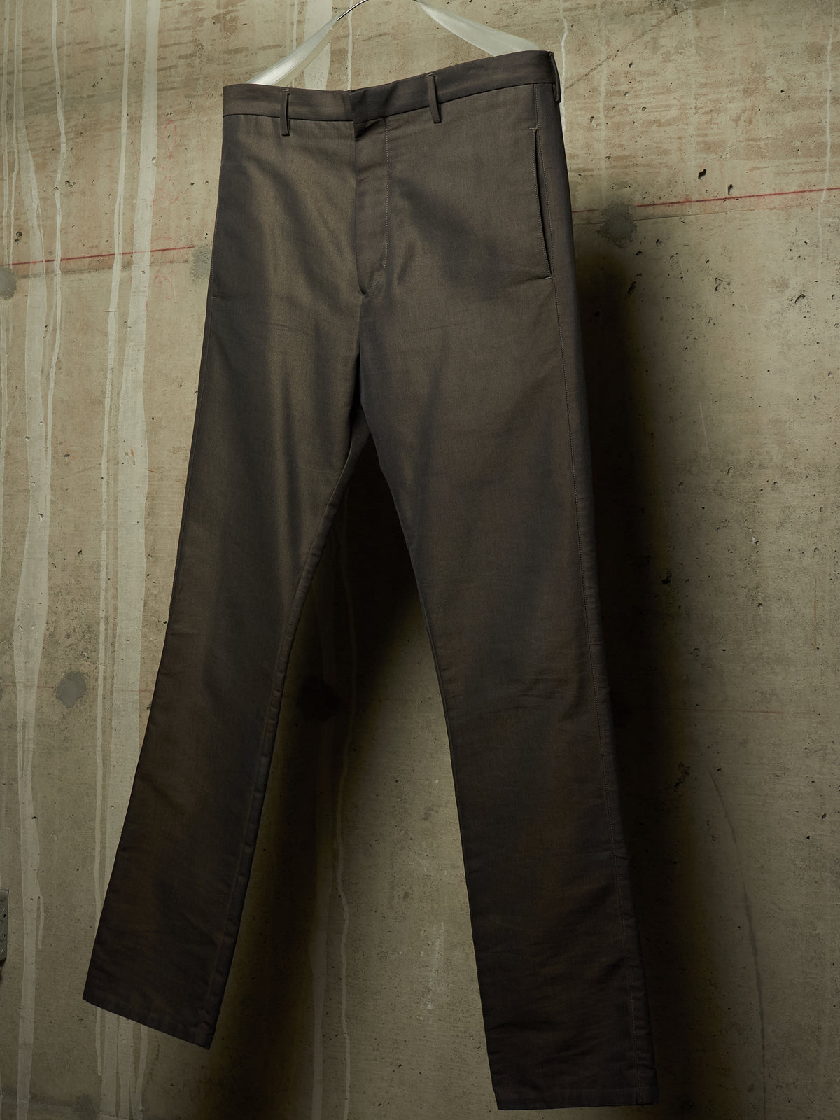 Dark brown slim-fit trousers hanging on a white plastic hanger against a textured concrete wall.