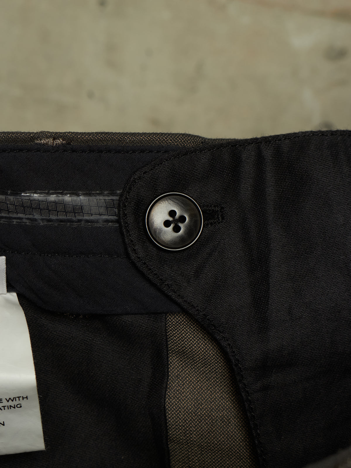 Close-up detail of black trousers showing a dark metal snap button, fabric texture, and a white size tag against a concrete background.