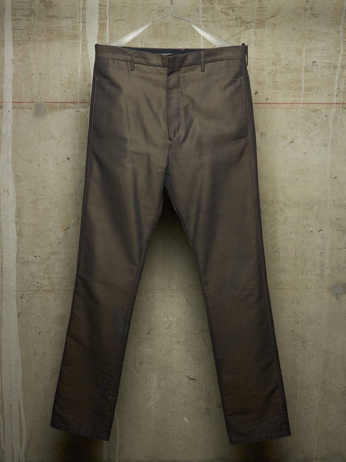 A pair of dark brown slim-fit trousers hanging from a white plastic hanger against a textured concrete wall.