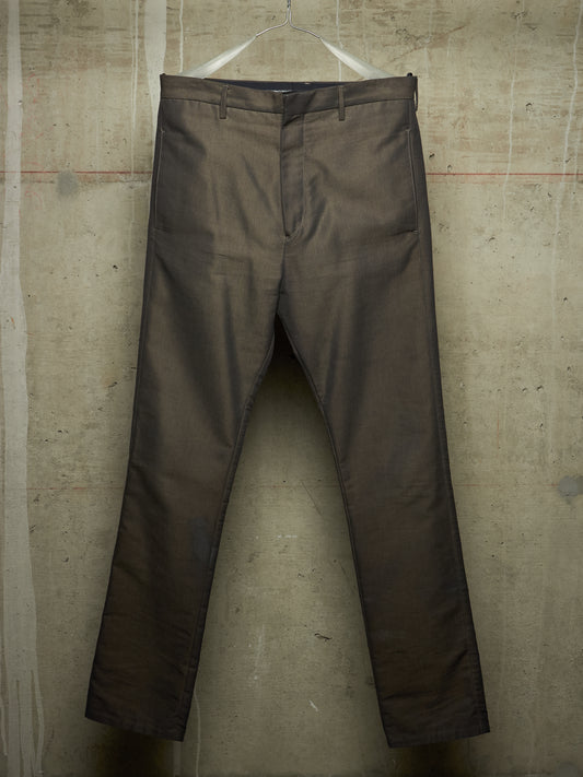 A pair of dark brown slim-fit trousers hanging from a white plastic hanger against a textured concrete wall.