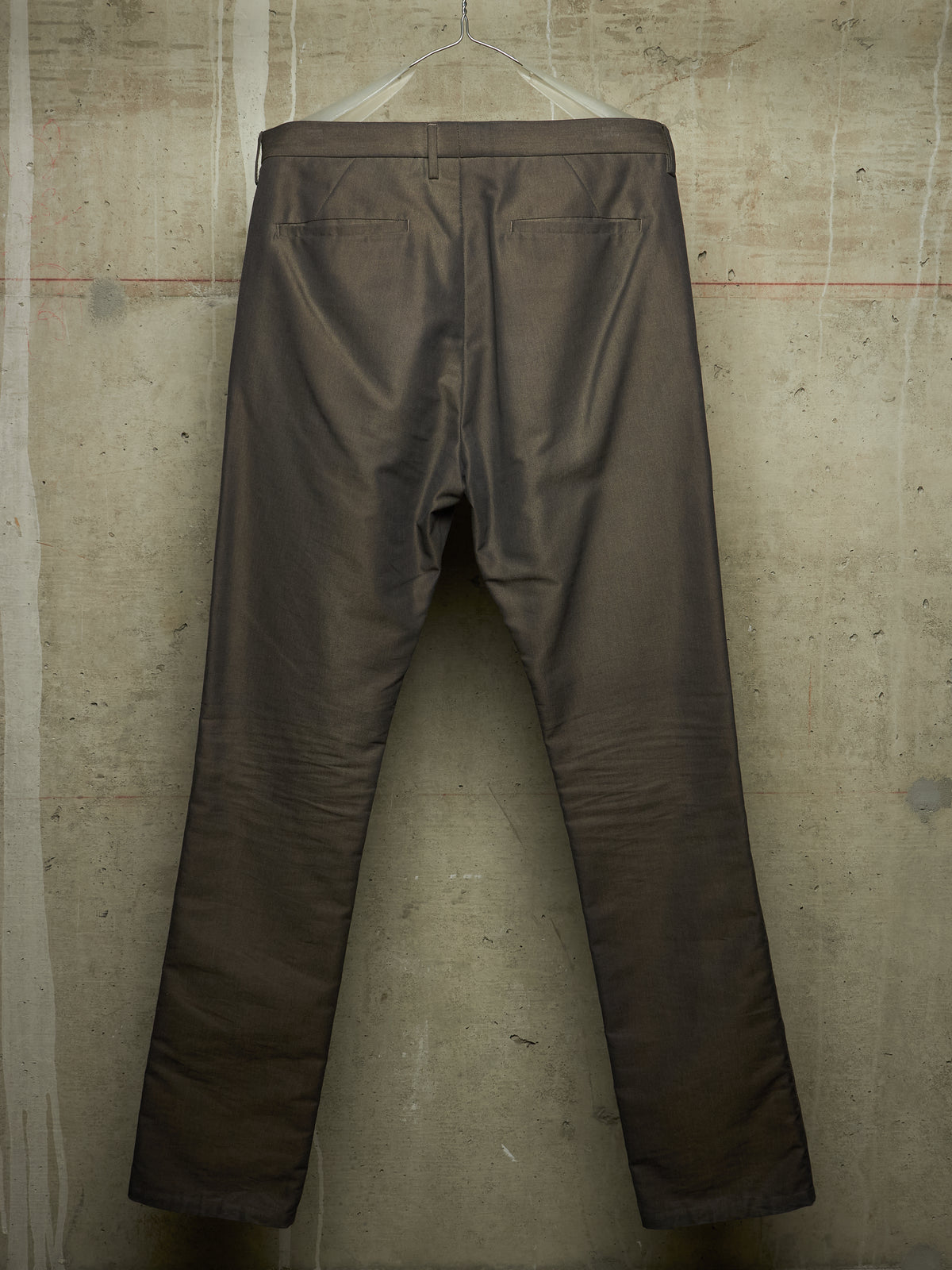 A pair of dark brown trousers hanging on a clear plastic hanger against a textured concrete wall.