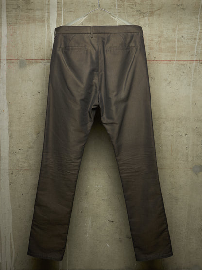 A pair of dark brown trousers hanging on a clear plastic hanger against a textured concrete wall.