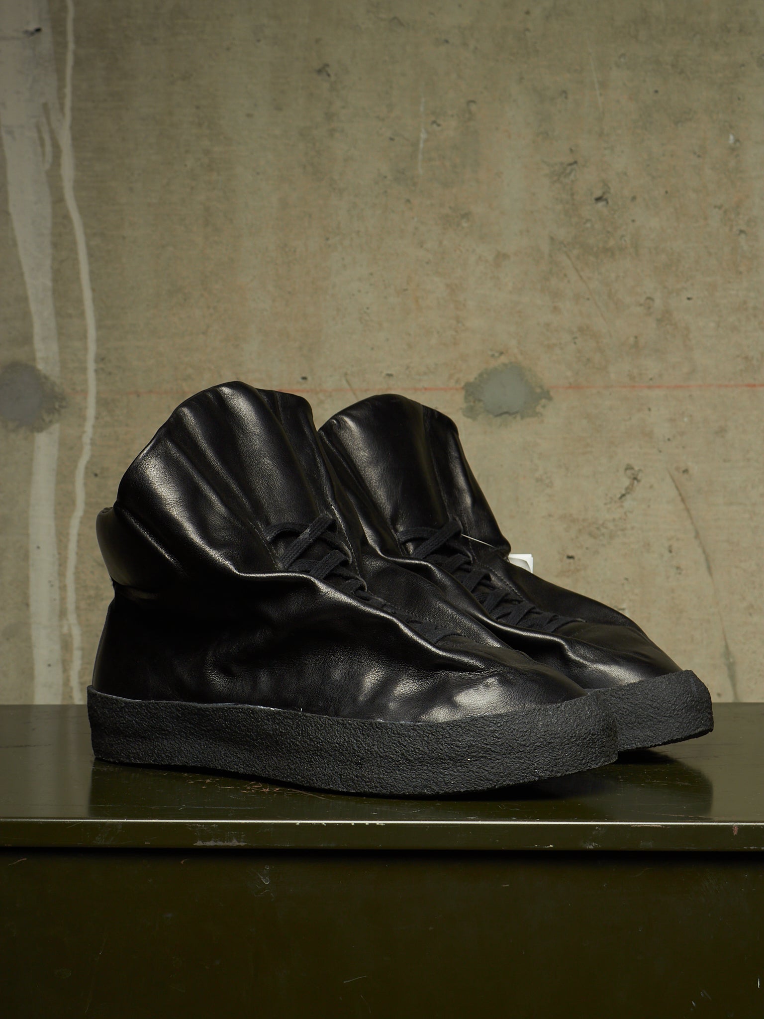 A pair of black leather high-top sneakers with sculpted, wrinkled textures and thick black soles, displayed on a dark green table against a textured concrete wall.